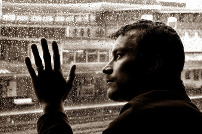A man looks out a window onto a cityscape. It is raining. The entire picture is in sepia tone.
